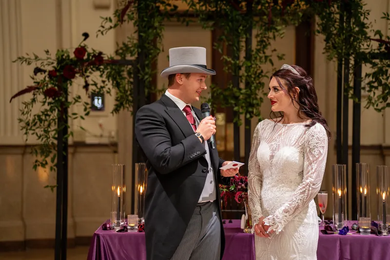 Sam speaks into a microphone while facing Alyson, with greenery and purple linens framing the reception space.