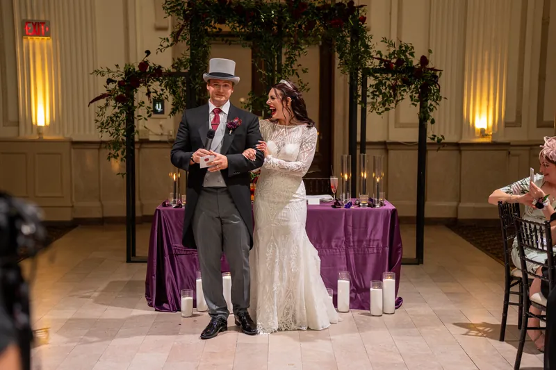 Sam and Alyson stand together on the reception floor as guests look on, with ivy garland and gold lighting behind them.