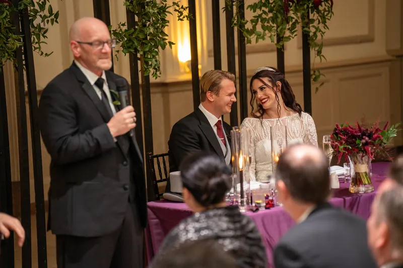 An officiant addresses guests as Sam and Alyson listen from their table with burgundy flowers and candles.
