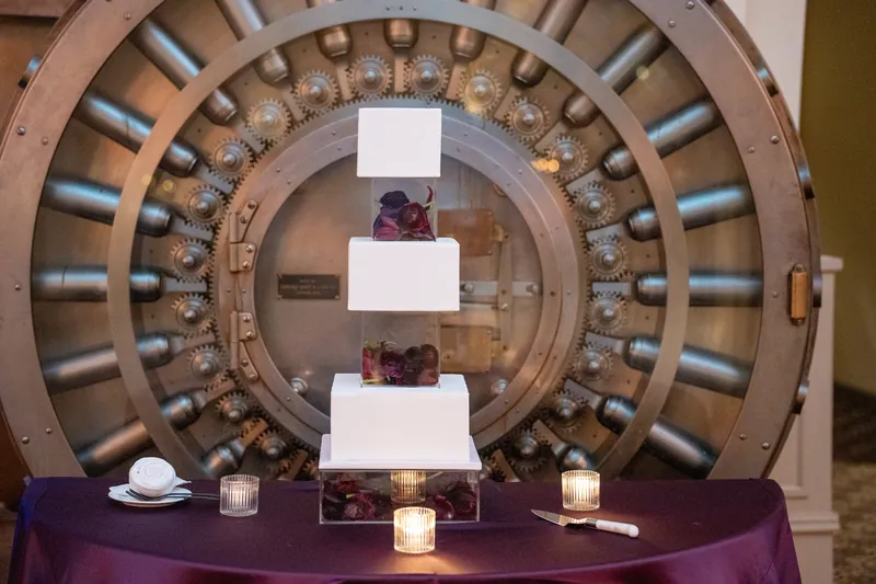 A three-tiered white cake sits beneath an ornate architectural dome, surrounded by candles and purple linens.