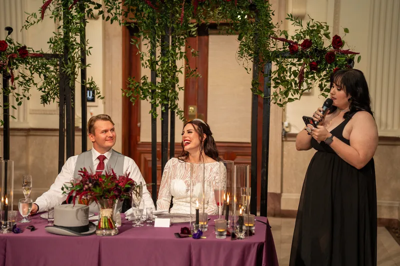 A woman in black speaks to guests while Sam and Alyson sit at their purple-draped table with flowers.