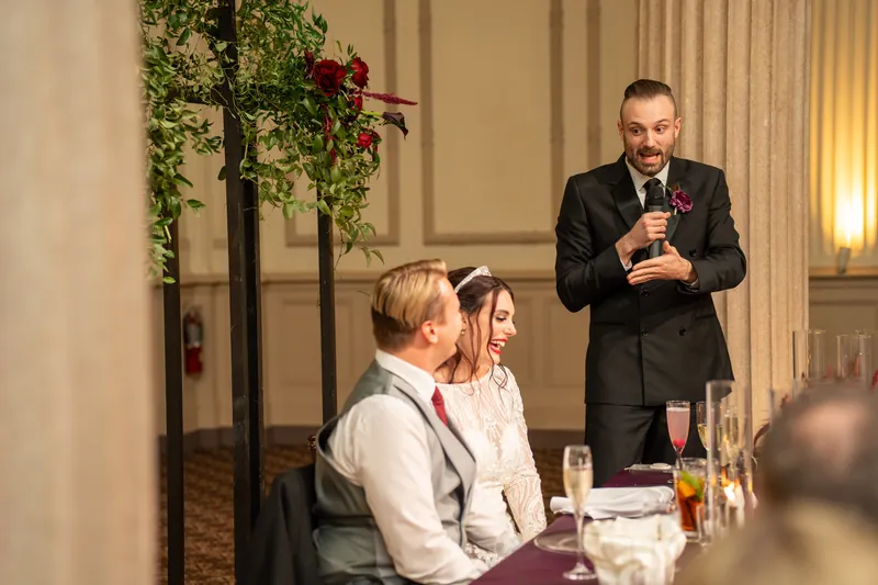 A groomsman raises his glass to speak as Sam and Alyson sit smiling at the reception table during toasts.