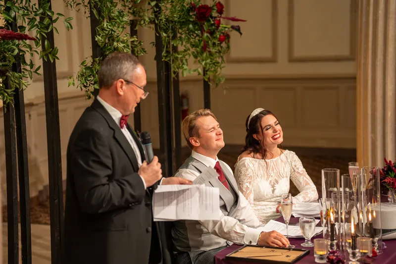 A man in a dark suit speaks at a reception table while Alyson and Sam listen from their seats at The Treasury on the Plaza.