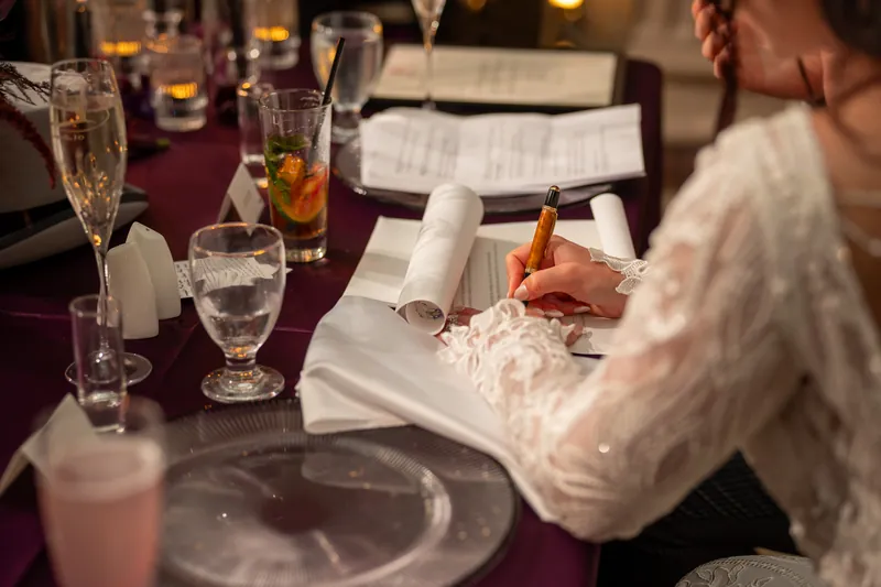 Someone in a cream lace sleeve signs a document at a reception table set with glasses and burgundy linens.