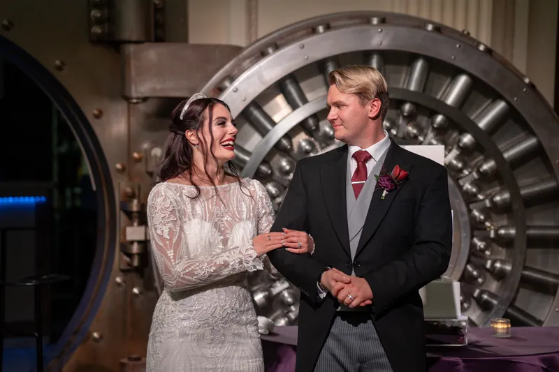 Alyson in a beaded lace gown and Sam in a charcoal suit hold hands and smile in front of the glowing gear art piece.