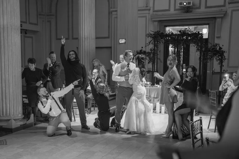 Alyson and Sam stand surrounded by guests in the elegant hall while someone adjusts their attire.