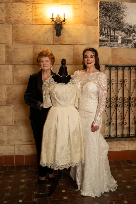 Alyson poses in a long lace gown with long sleeves while another woman in a dark jacket holds a black clutch beside her.