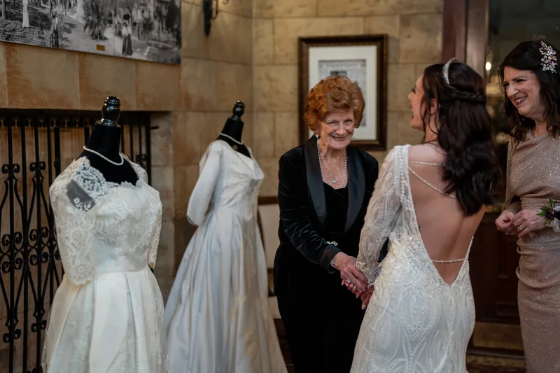 A woman with red hair chats with Alyson, who wears a backless white gown, as another guest looks on nearby.
