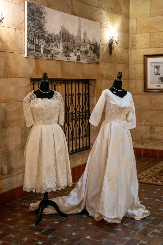 Two white lace wedding gowns displayed on dress forms hang against a stone wall with vintage photographs above.