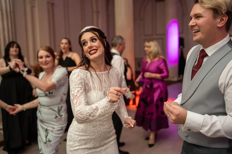 Alyson and Sam laugh together on the dance floor, holding hands while other guests celebrate around them.