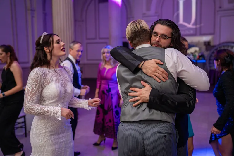 Sam hugs a guest wearing a gray sweater while Alyson smiles nearby in her white lace gown during the reception celebration.