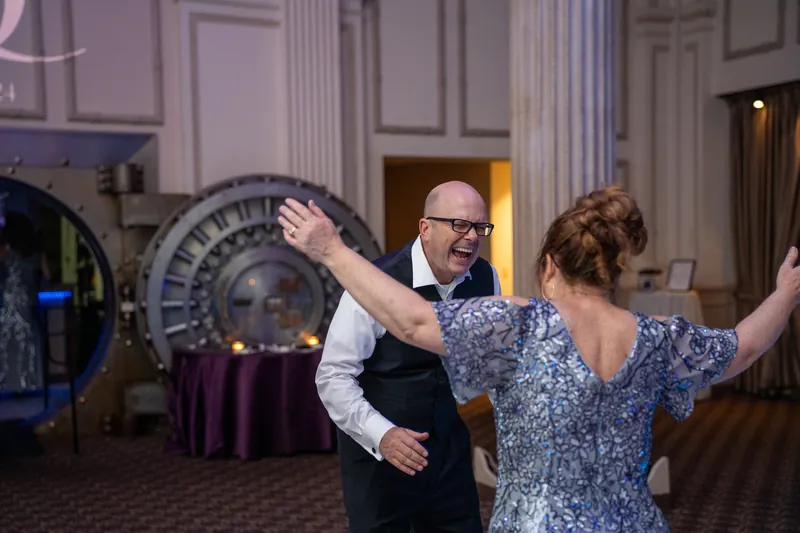 Sam dances with arms outstretched while a woman in a blue patterned dress celebrates beside him at the ballroom gathering.