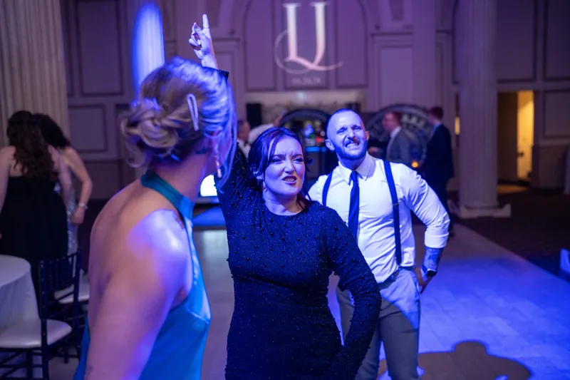 Guests dance under blue lighting, including a woman in a dark blue dress smiling as the party continues around her.