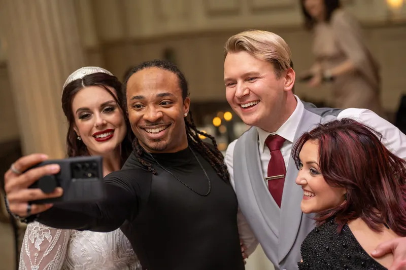 Four wedding guests smile together as one holds up a phone to capture a selfie moment.