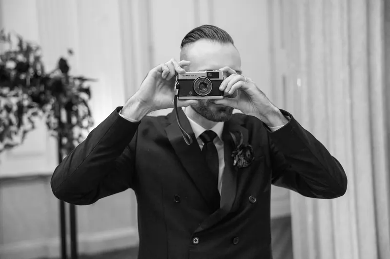 A guest in a black suit and red tie raises a vintage camera to his face, snapping a photo during the reception.