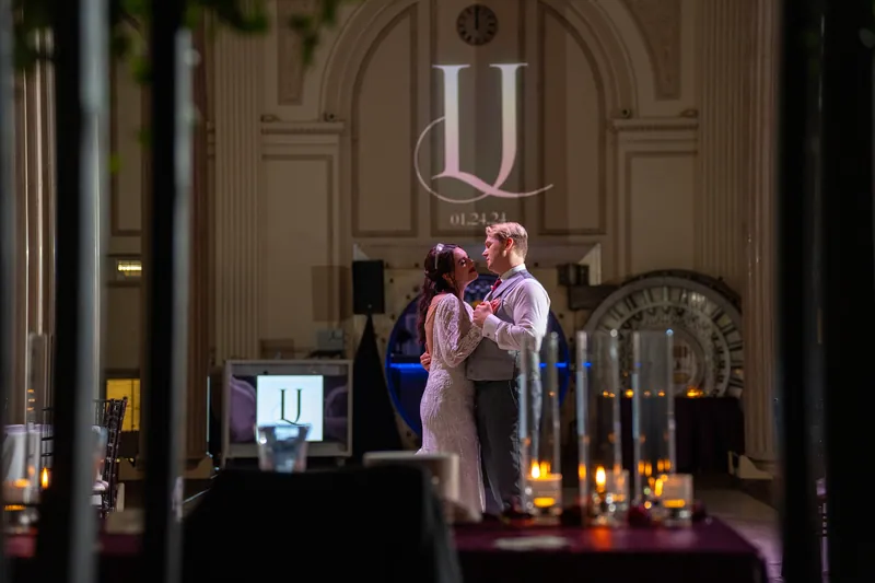 Alyson and Sam stand at the altar beneath a glowing monogram in candlelit architectural grandeur.