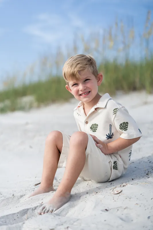 A young boy in a patterned shirt sits smiling on the sand at Vilano Beach with beach grass and dunes behind him.
