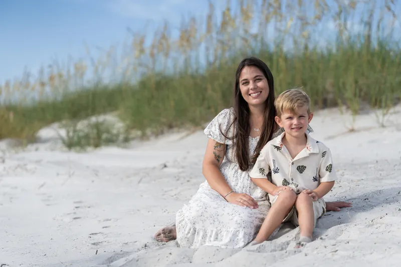 Ana and a young boy sit together on the sand at Vilano Beach, both smiling at the camera with dune vegetation