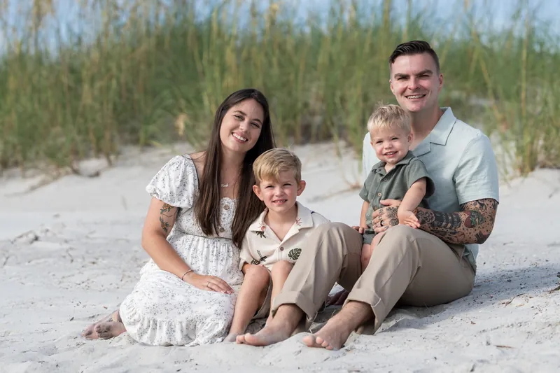 Ana, Bailey, and their two children sit together on the sand at Vilano Beach with dunes and beach grass visible behind them.