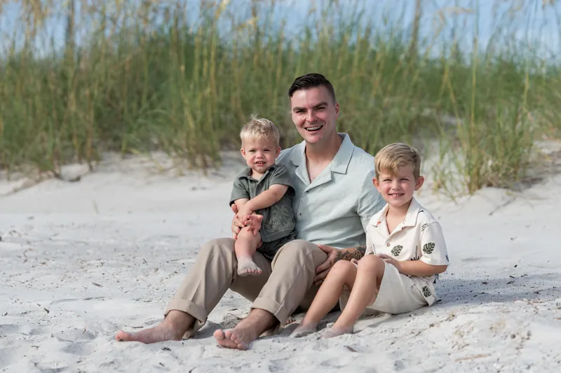 Bailey holds a toddler while sitting on the sand with two other children at Vilano Beach on a sunny day.