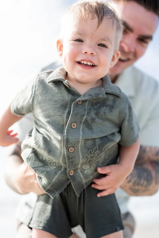 A young child in a gray button-up shirt grins at the camera while being held by Bailey at the beach.