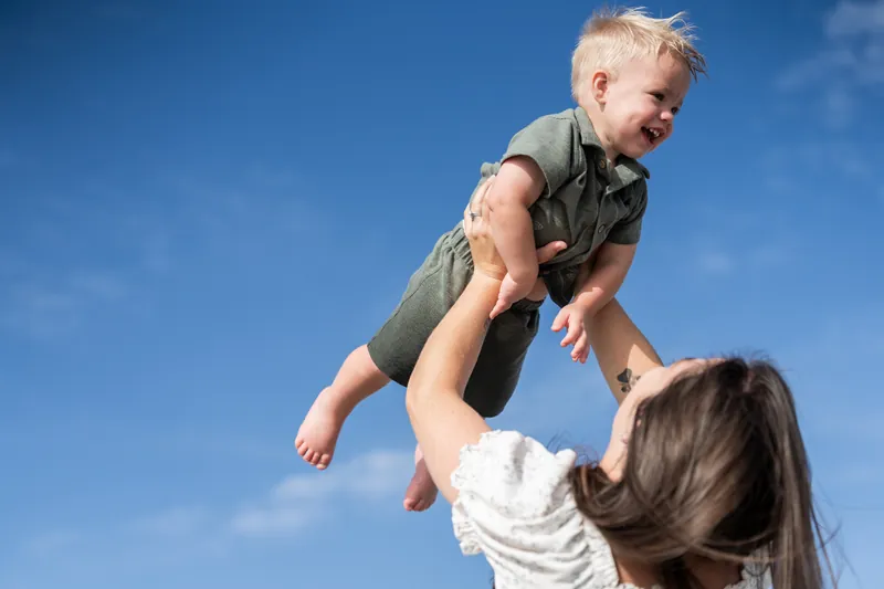 Bailey lifts their smiling child high against a bright blue sky at Vilano Beach, St. Augustine.