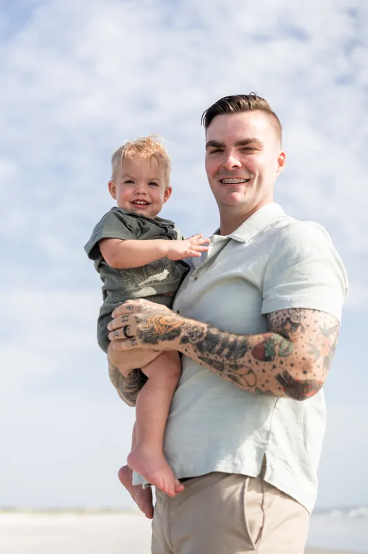 Bailey holds their toddler, both smiling, with the beach and cloudy sky visible behind them at Vilano Beach, St. Augustine.