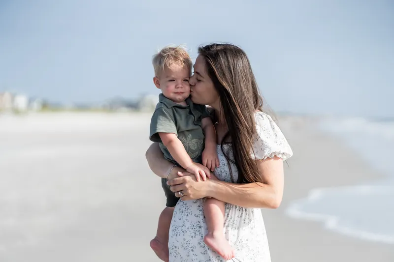 Ana carries their young child and kisses their cheek while standing on the sand at Vilano Beach, St. Augustine.