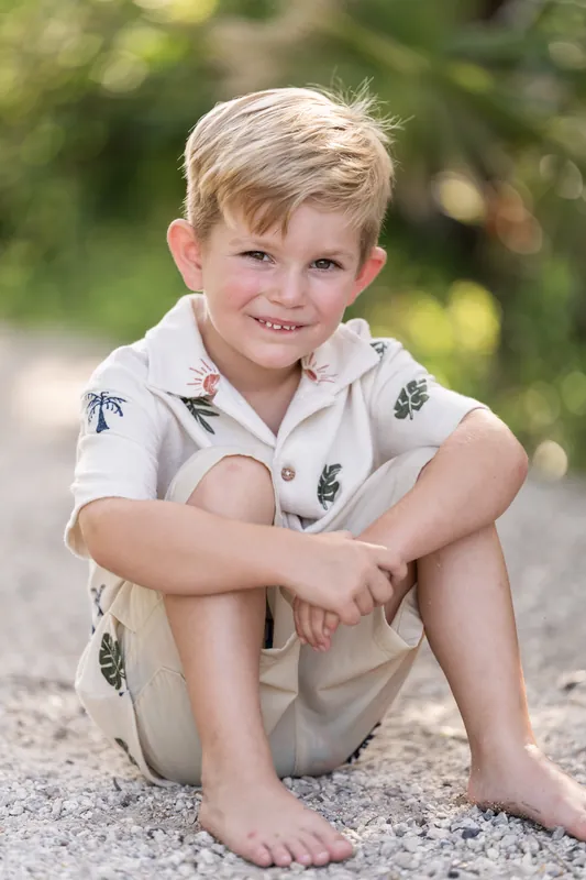 A young child in a patterned shirt and khaki shorts sits on a gravel path, smiling with arms crossed.