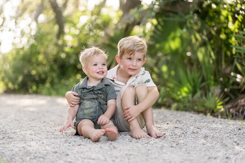 Two young children sit barefoot together on a sandy path surrounded by green foliage, looking at each other.