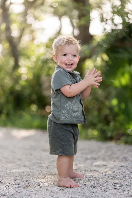 A toddler in a gray patterned shirt stands on a path with arms at their sides, beaming with a big smile.