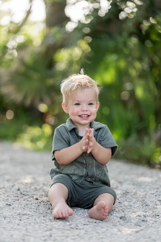 A small child sits on gravel wearing a gray button-up shirt, clasping their hands together with a joyful expression.