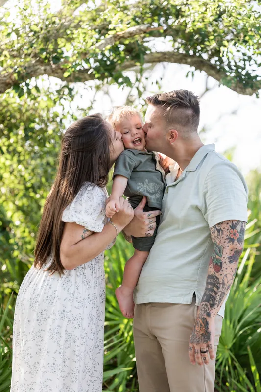 Ana and Bailey hold their toddler together under flowering vines, smiling at their child in a lush garden setting.