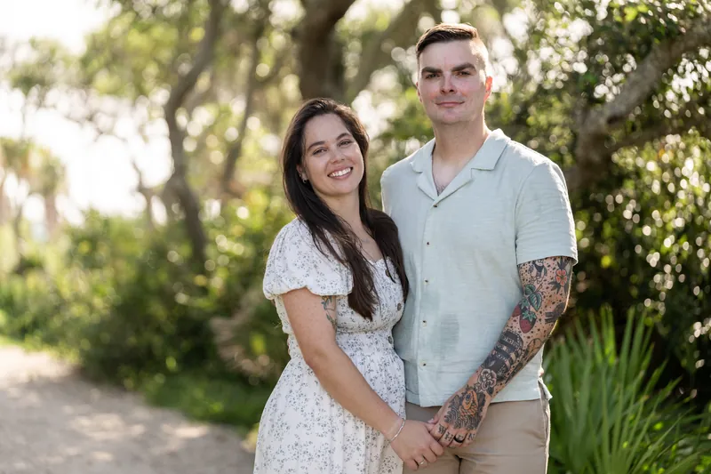 Ana and Bailey stand close together on a tree-lined path, holding hands and looking at the camera with soft smiles.