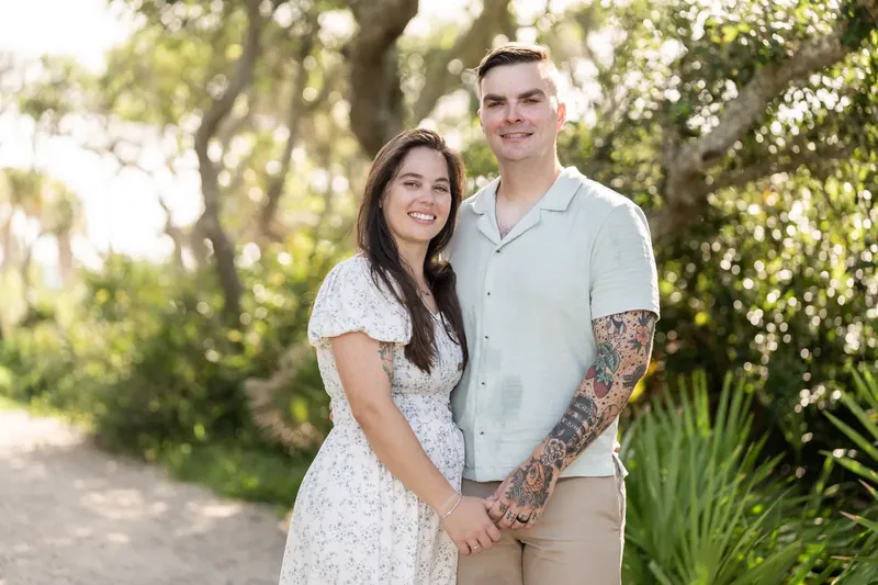 The couple walks hand in hand down a tree-lined path at the beachfront park, with lush greenery surrounding them.