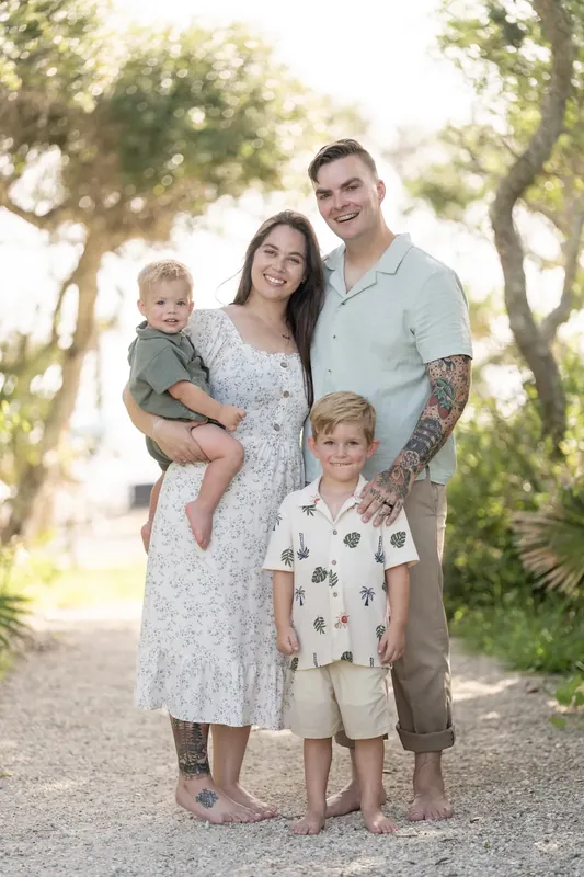 Ana and Bailey stand together on a shaded path with two children, all facing the camera in casual summer clothing.