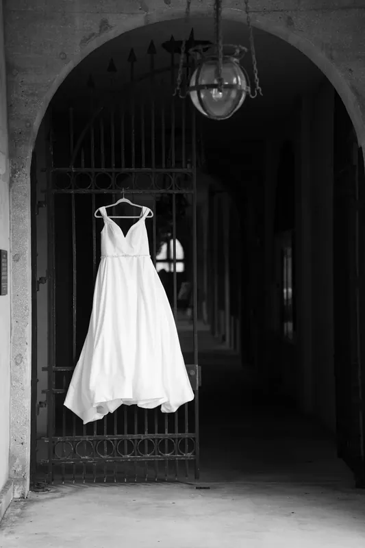 White wedding dress hangs from an ornate metal gate beneath a pendant light in a dramatic archway at Lightner Museum.