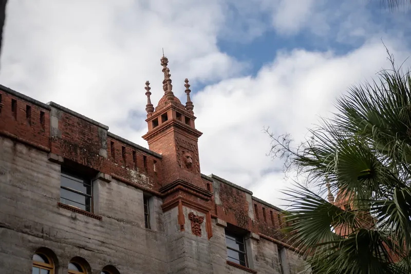 Ornate brick tower with decorative spires rises above the roofline of Lightner Museum under a partly cloudy sky.