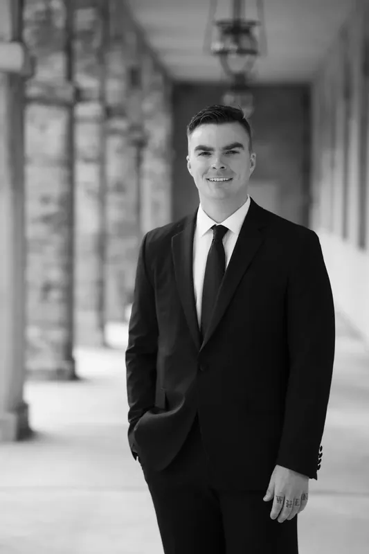Bailey stands in a black suit and tie in a columned corridor, smiling at the camera.