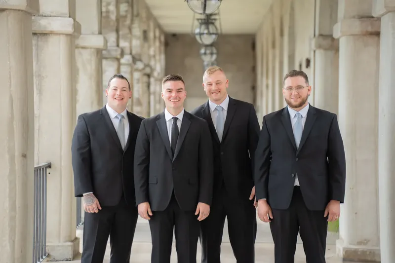 Four people in dark suits stand shoulder-to-shoulder in a classical colonnade at Lightner Museum.