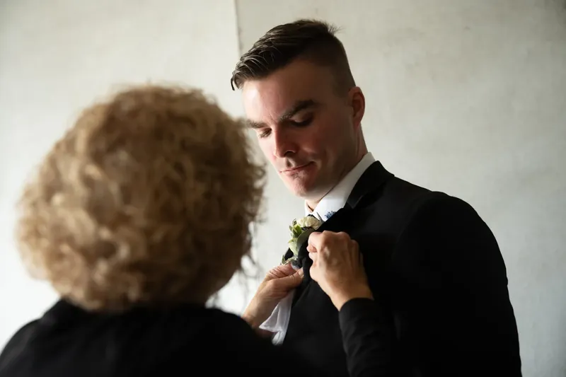 Someone adjusts a boutonnière on Bailey's chest as he looks down with a slight smile.