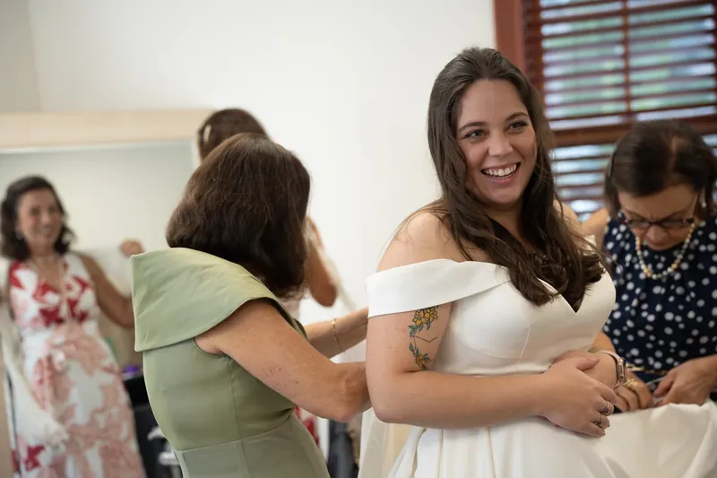 Bailey smiles while loved ones help adjust her white off-shoulder wedding dress at Amore Wedding Chapel.