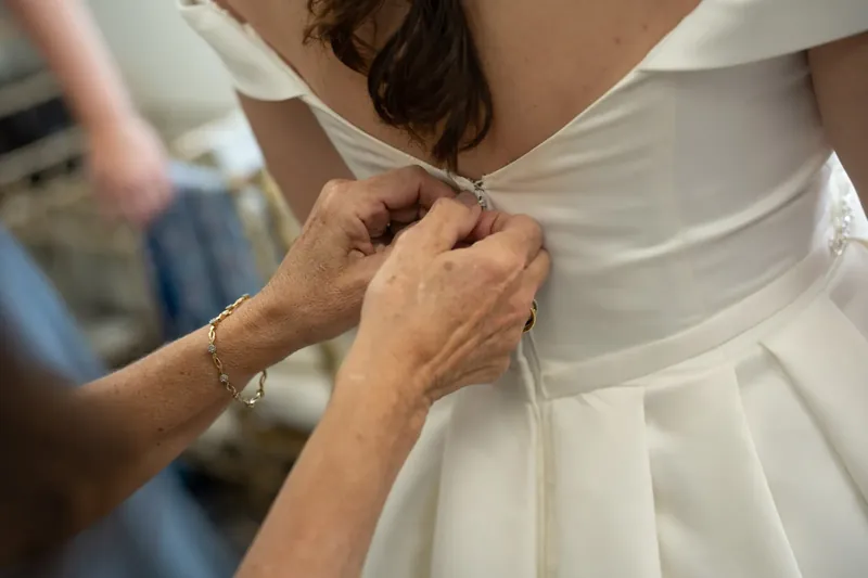 Hands fasten the back of Bailey's white gown as she prepares for her ceremony.