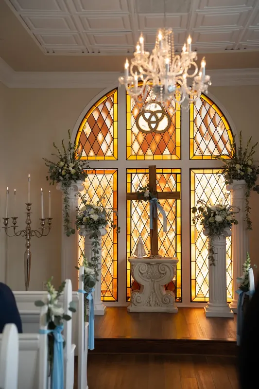 An ornate arched stained-glass window glows warmly above the altar at Amore Wedding Chapel, flanked by floral arrangements.