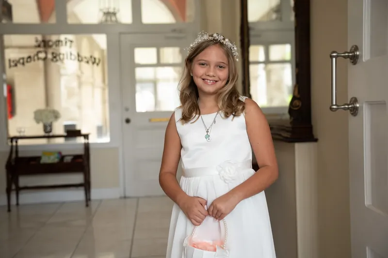 A young attendant in a white dress and tiara smiles with clasped hands at Amore Wedding Chapel.