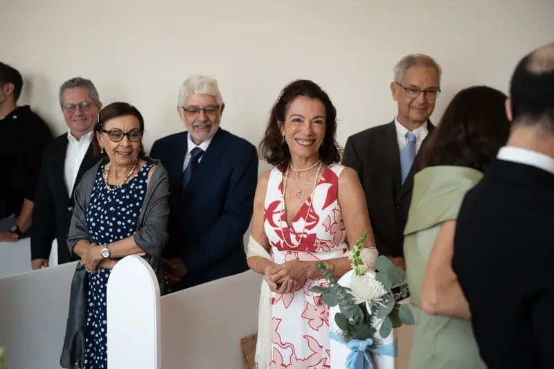 Wedding guests in formal attire gather at the chapel, a woman in a red floral dress holding a white bouquet.