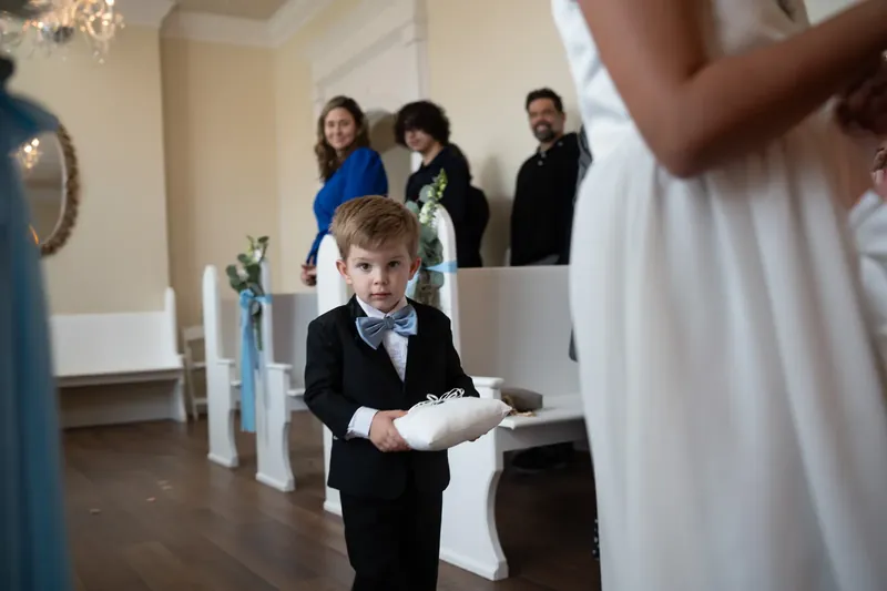 A young ring bearer in a black suit carries a white pillow down the chapel aisle as guests watch.