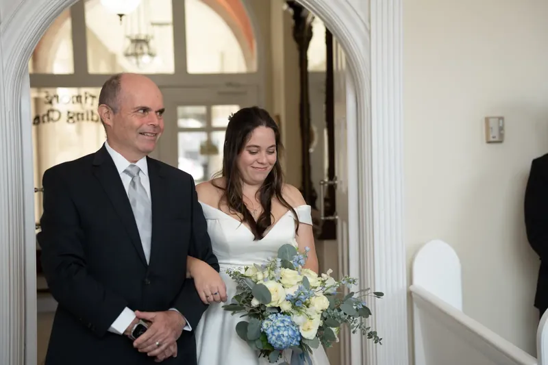 Ana and Bailey walk together through an arched doorway, Ana in white and Bailey in a dark suit.