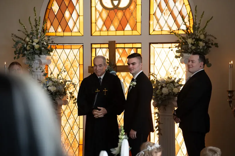 Ana and Bailey stand before a stained-glass window with floral arrangements during their ceremony.