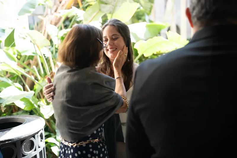 Ana greets a guest outdoors at Lightner Museum, smiling while shaking hands surrounded by lush green foliage.